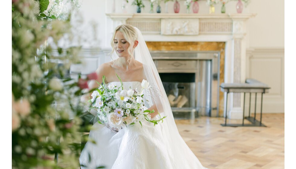 Bride with side-swept curls and veil holds a bouquet in country house lounge before her Hampshire garden wedding.